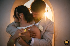 Bride and groom hugging indoors at sunset in Santorini, Greece, with sunlight bursting through the window behind them, creating an intimate atmosphere.